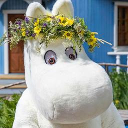 A Moomin character stands in front of the blue Moominhouse at Moominworld.